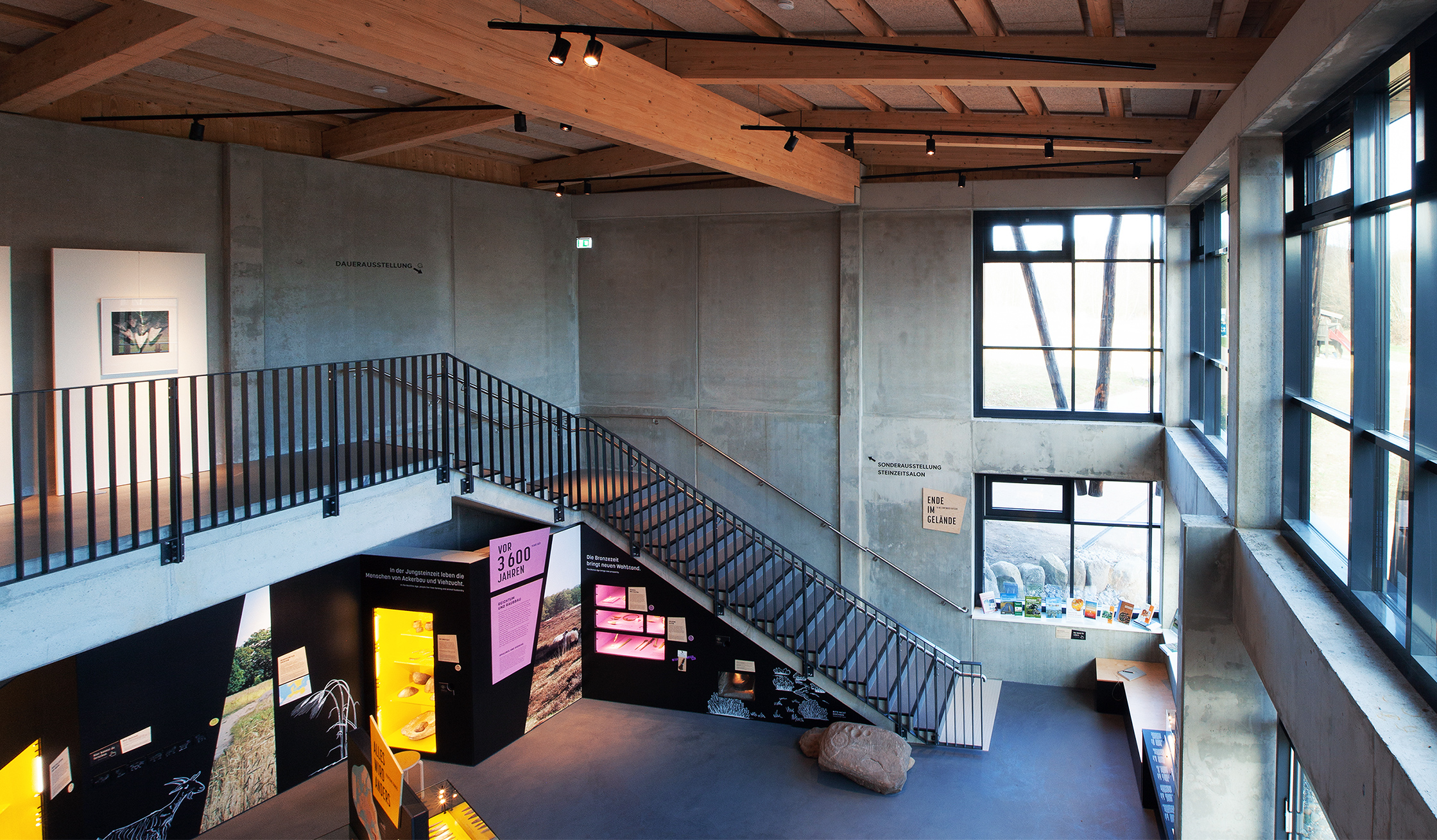 Referenz Steinzeithaus Albersdorf: Großzügiger Ausstellungsraum mit Betonwänden, viel Glas und einer Treppe. Unten sind bunte Informationstafeln und Vitrinen mit Exponaten zu sehen.