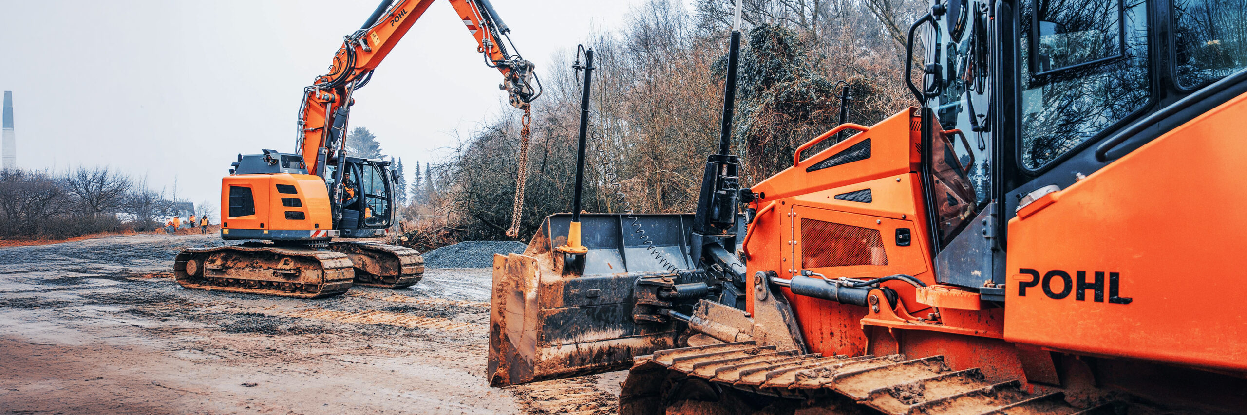Zwei Baumaschinen von POHL auf einer matschigen Baustelle, einer hebt Material mit Kette an.