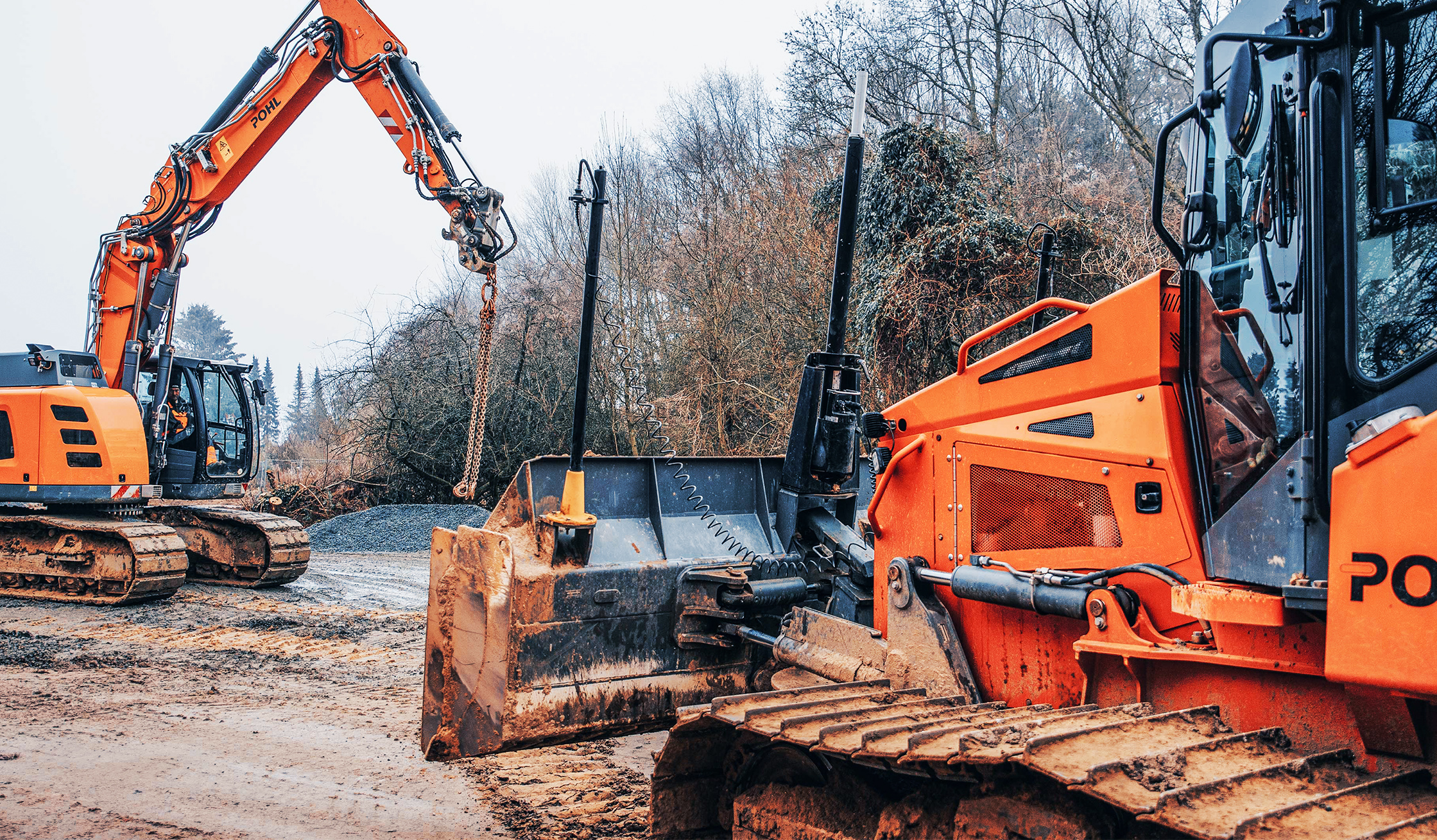 Zwei Baumaschinen von POHL auf einer matschigen Baustelle, einer hebt Material mit Kette an.