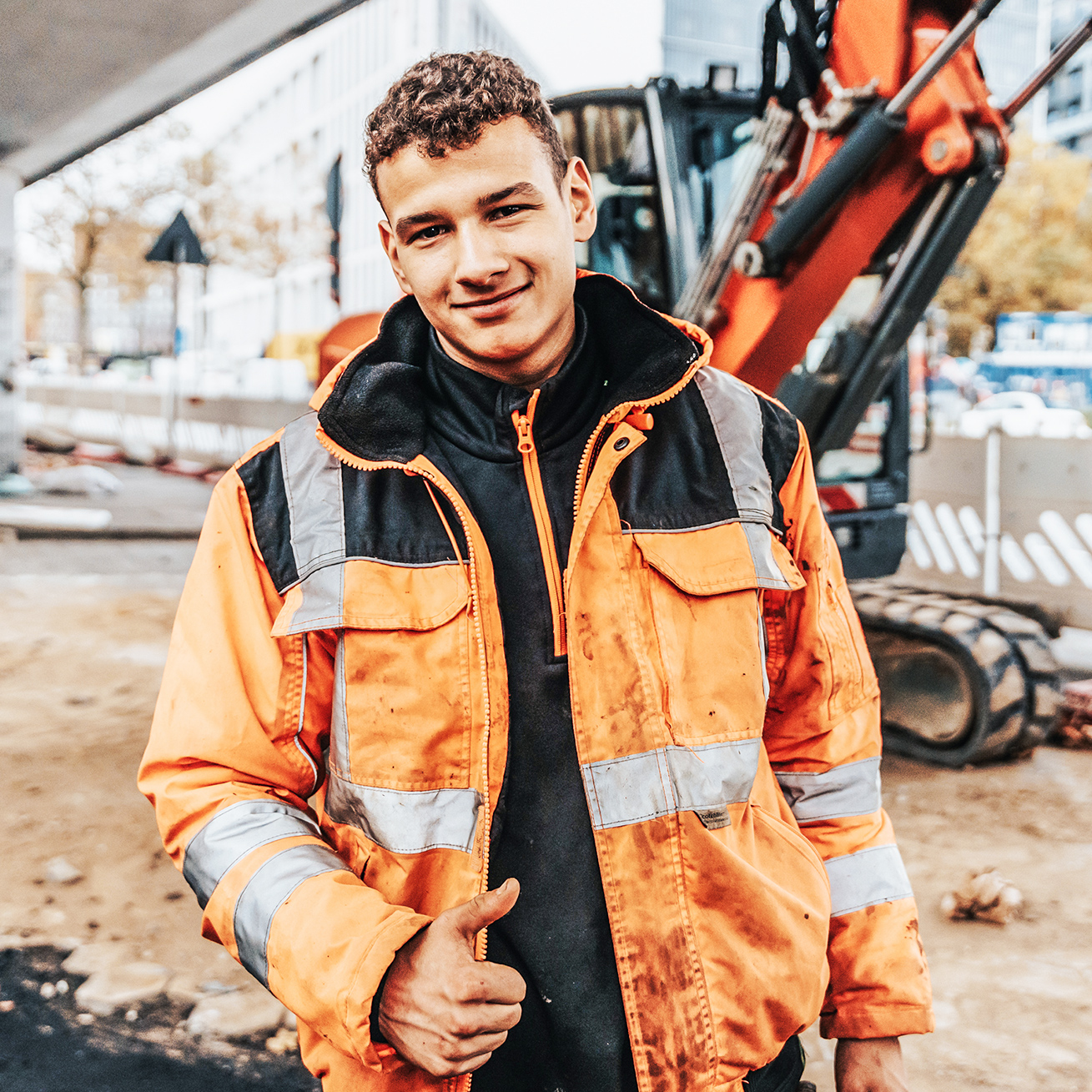 In der Mitte steht ein junger POHL-Mitarbeiter in orange Jacke auf einer innerstädtischen Baustelle und hält den Daumen hoch. Links hinten steht ein orange POHL-Bagger; rechts begrenzen weiße Absperrelemente die Fahrbahn.