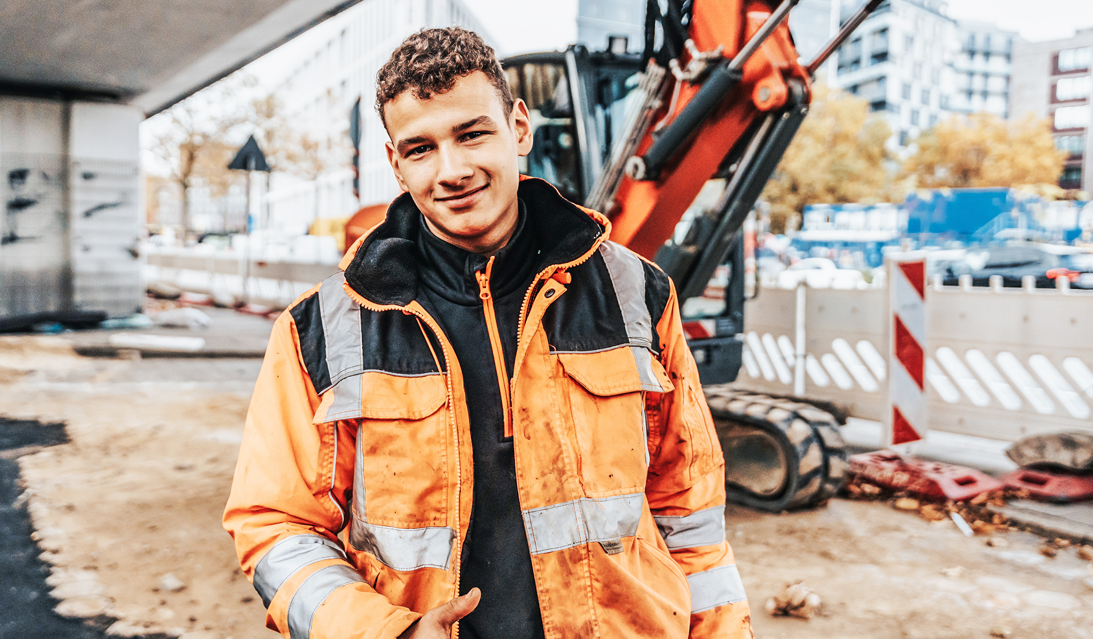 In der Mitte steht ein junger POHL-Mitarbeiter in orange Jacke auf einer innerstädtischen Baustelle und hält den Daumen hoch. Links hinten steht ein orange POHL-Bagger; rechts begrenzen weiße Absperrelemente die Fahrbahn.