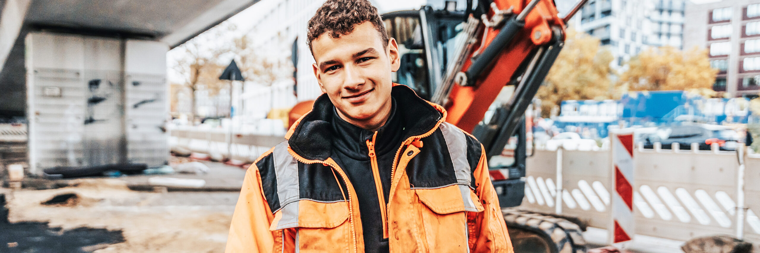 In der Mitte steht ein junger POHL-Mitarbeiter in orange Jacke auf einer innerstädtischen Baustelle und hält den Daumen hoch. Links hinten steht ein orange POHL-Bagger; rechts begrenzen weiße Absperrelemente die Fahrbahn.
