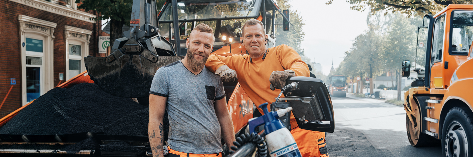 In der Mitte lehnen zwei Männer an einer großen Straßenbaumaschine mit gefüllter Schaufel. Rechts im Bild steht ein orangefarbenes POHL-Fahrzeug am Fahrbahnrand. Links im Bild sind die Fassaden eines Backsteingebäudes. Vorne im Bild liegt ein Haufen dunkles Material. Im Hintergrund verläuft eine Straße mit Bäumen und weiteren Fahrzeugen.
