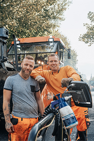 In der Mitte lehnen zwei Männer an einer großen Straßenbaumaschine mit gefüllter Schaufel. Rechts im Bild steht ein orangefarbenes POHL-Fahrzeug am Fahrbahnrand. Links im Bild sind die Fassaden eines Backsteingebäudes. Vorne im Bild liegt ein Haufen dunkles Material. Im Hintergrund verläuft eine Straße mit Bäumen und weiteren Fahrzeugen.