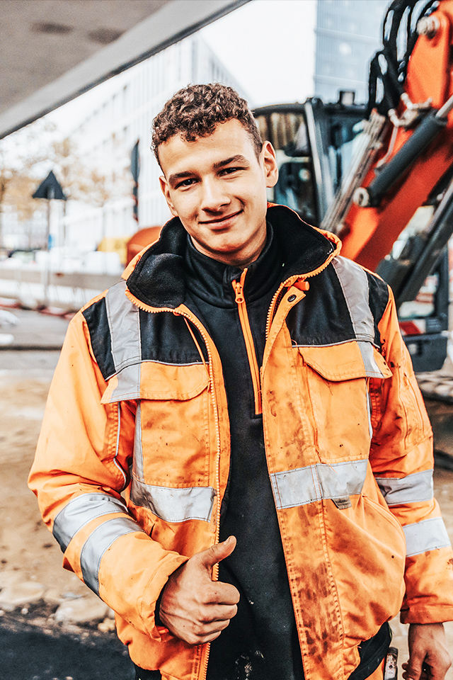 In der Mitte steht ein junger POHL-Mitarbeiter in orange Jacke auf einer innerstädtischen Baustelle und hält den Daumen hoch. Links hinten steht ein orange POHL-Bagger; rechts begrenzen weiße Absperrelemente die Fahrbahn.