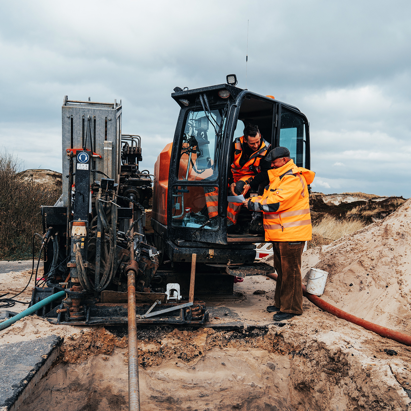 Standort Sylt: In der Mitte steht eine orange POHL-Maschine am Rand eines Sandhaufens. Links daneben sprechen zwei POHL-Mitarbeiter in orangefarbener Kleidung; rechts im Hintergrund liegen Dünen und Büsche.