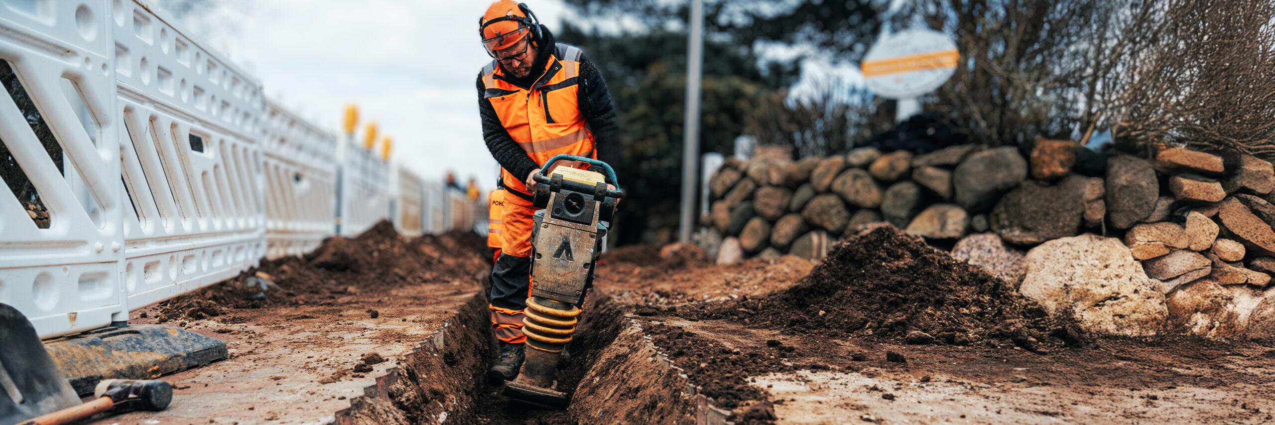 Ein Arbeiter in kompletter POHL-Schutzkleidung mit Helm arbeitet tief in einem schmalen Graben. Er hält eine vibrierende Maschine, die den braunen Boden verdichtet. Weiße Absperrgitter und ein Verkehrsschild stehen im Hintergrund.