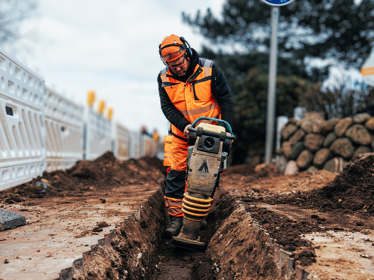 Ein Arbeiter in kompletter POHL-Schutzkleidung mit Helm arbeitet tief in einem schmalen Graben. Er hält eine vibrierende Maschine, die den braunen Boden verdichtet. Weiße Absperrgitter und ein Verkehrsschild stehen im Hintergrund.