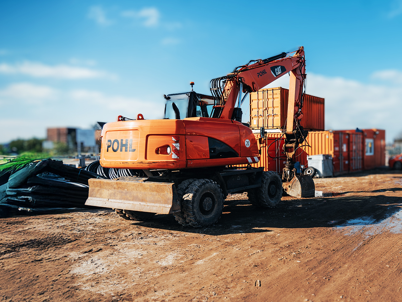 Ein riesiger orangefarbener POHL-Bagger parkt auf einer Baustelle. Neben ihm stehen Container, der Boden ist uneben und voller Sand und Steine.