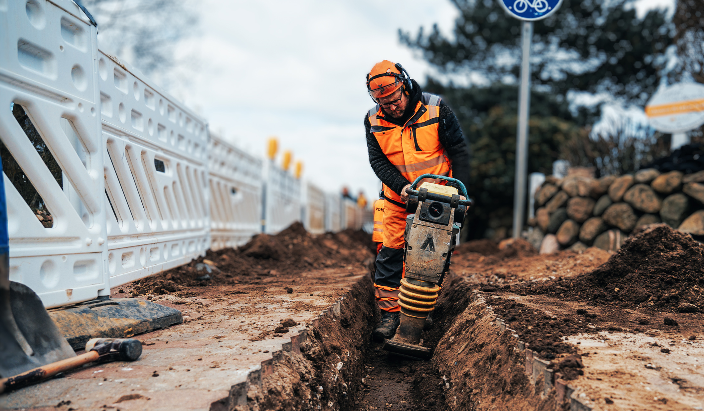 Ein Arbeiter in kompletter POHL-Schutzkleidung mit Helm arbeitet tief in einem schmalen Graben. Er hält eine vibrierende Maschine, die den braunen Boden verdichtet. Weiße Absperrgitter und ein Verkehrsschild stehen im Hintergrund.