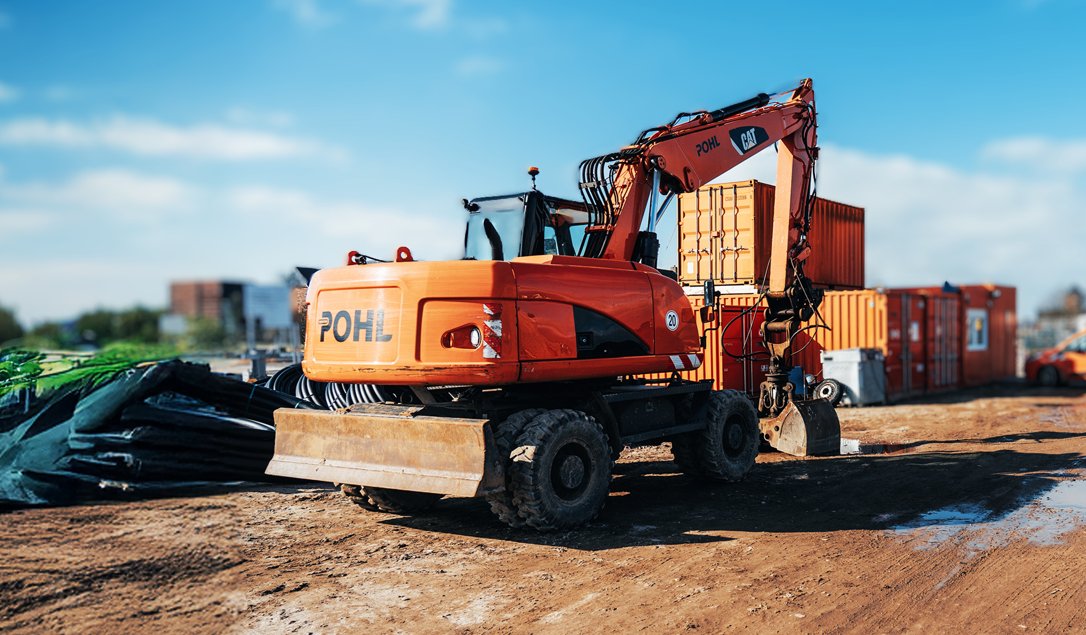 Referenz Sylt Terminalfläche: Ein riesiger orangefarbener POHL-Bagger parkt auf einer Baustelle. Neben ihm stehen Container, der Boden ist uneben und voller Sand und Steine.
