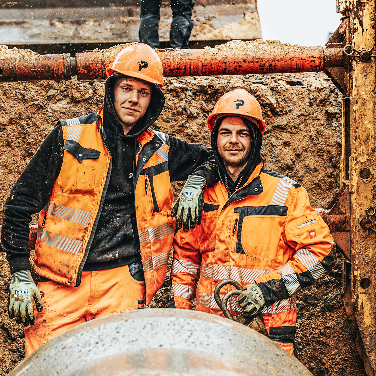 In der Mitte stehen zwei POHL-Mitarbeiter in orange Schutzkleidung und Helmen in einer Baugrube. Links und rechts halten Holzbohlenwände und rote Stahlstreben die Grube; vorne unten liegt ein rundes Rohr.