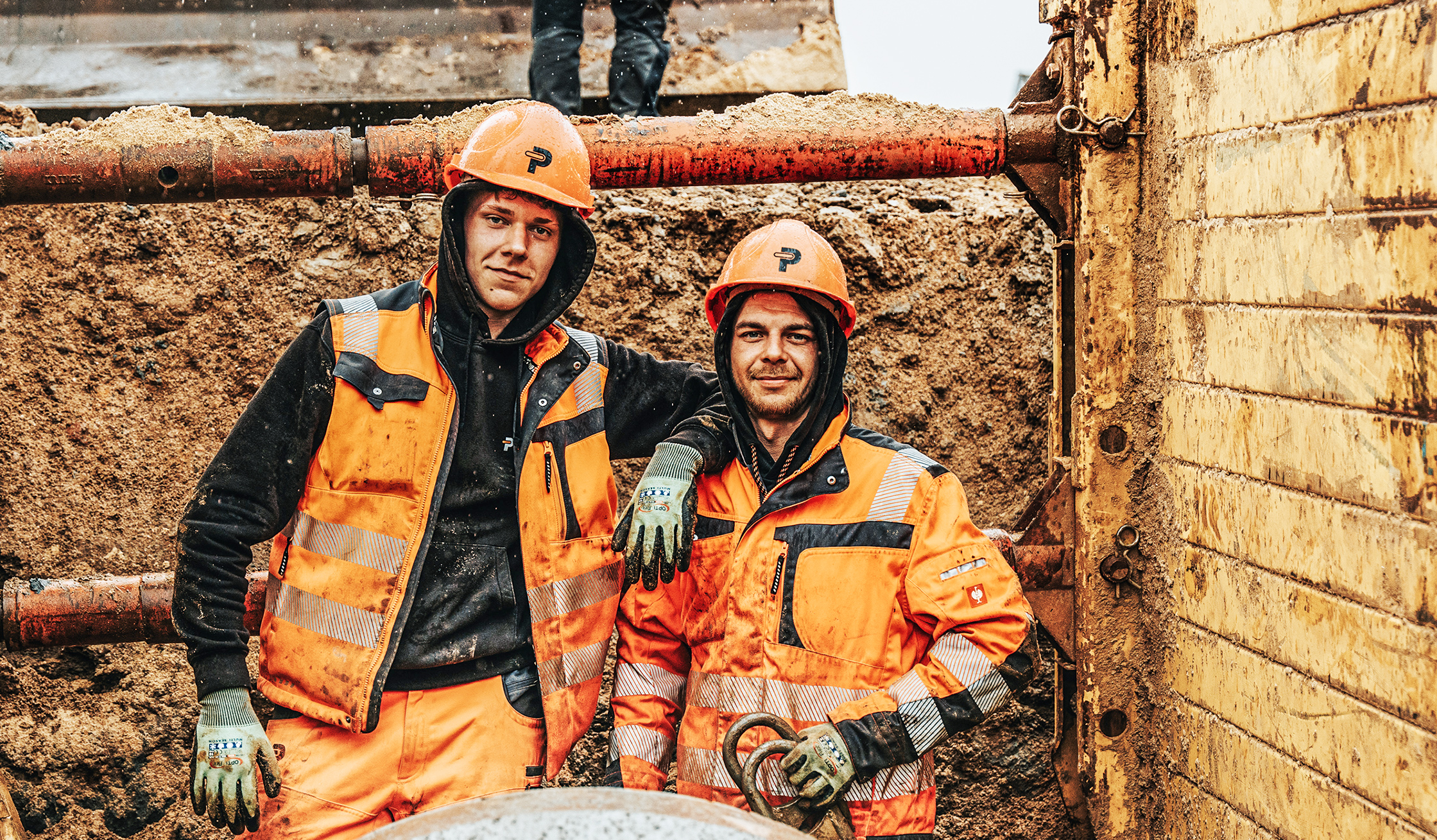 In der Mitte stehen zwei POHL-Mitarbeiter in orange Schutzkleidung und Helmen in einer Baugrube. Links und rechts halten Holzbohlenwände und rote Stahlstreben die Grube; vorne unten liegt ein rundes Rohr.
