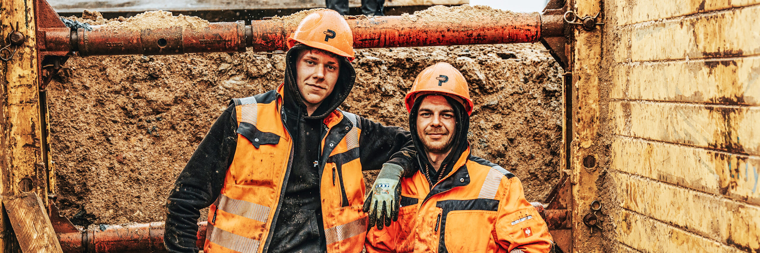 In der Mitte stehen zwei POHL-Mitarbeiter in orange Schutzkleidung und Helmen in einer Baugrube. Links und rechts halten Holzbohlenwände und rote Stahlstreben die Grube; vorne unten liegt ein rundes Rohr.