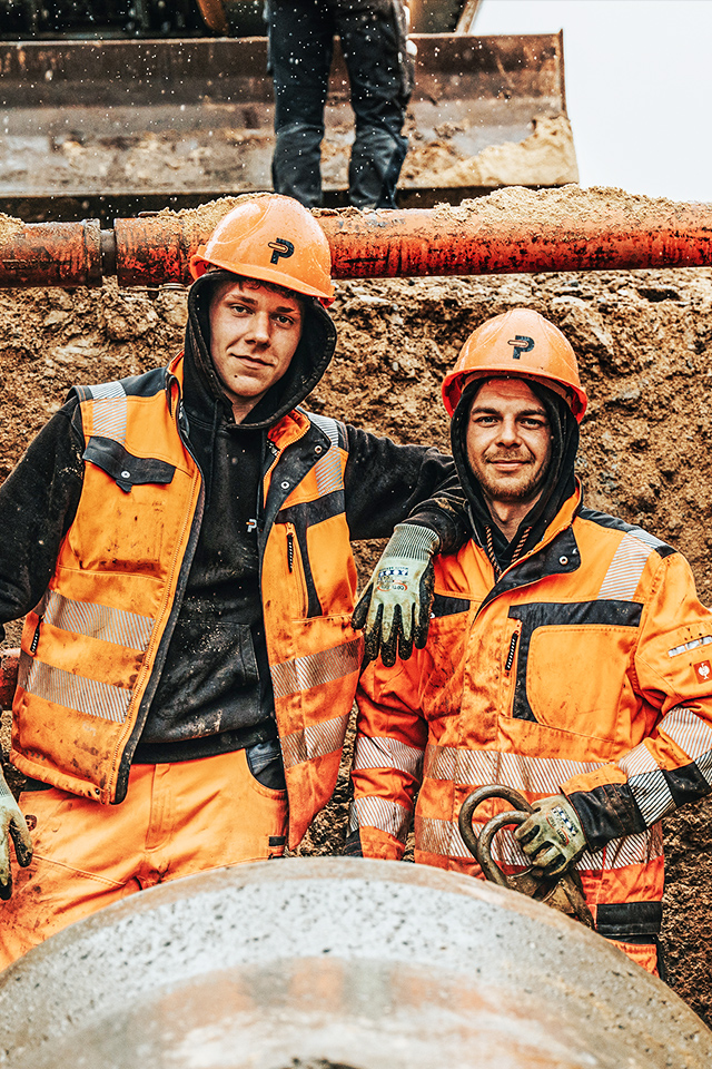In der Mitte stehen zwei POHL-Mitarbeiter in orange Schutzkleidung und Helmen in einer Baugrube. Links und rechts halten Holzbohlenwände und rote Stahlstreben die Grube; vorne unten liegt ein rundes Rohr.