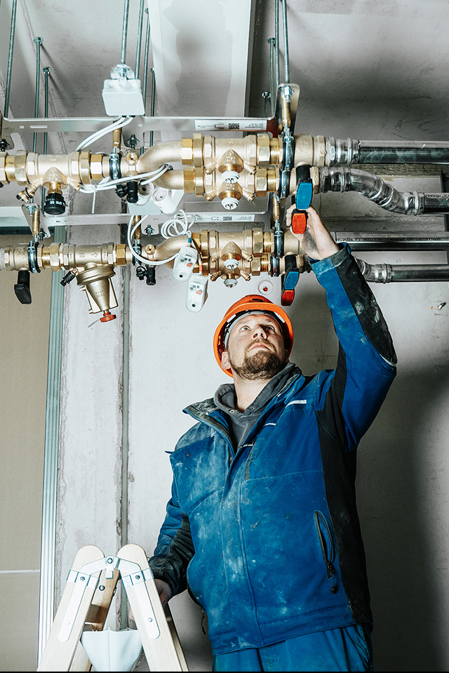 POHL Handwerker in blauer Schutzkleidung und Helm montiert Rohre und Ventile an der Decke eines Gebäudes.