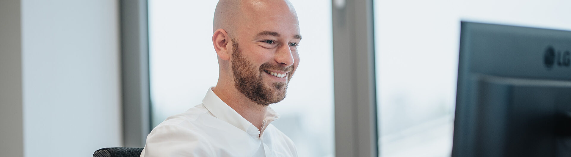 In der Mitte sitzt eine Person im weißen Hemd an einem Schreibtisch und schaut auf einen Monitor; links im Hintergrund steht ein großes Fenster.