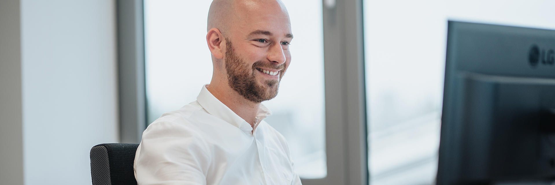 In der Mitte sitzt eine Person im weißen Hemd an einem Schreibtisch und schaut auf einen Monitor; links im Hintergrund steht ein großes Fenster.