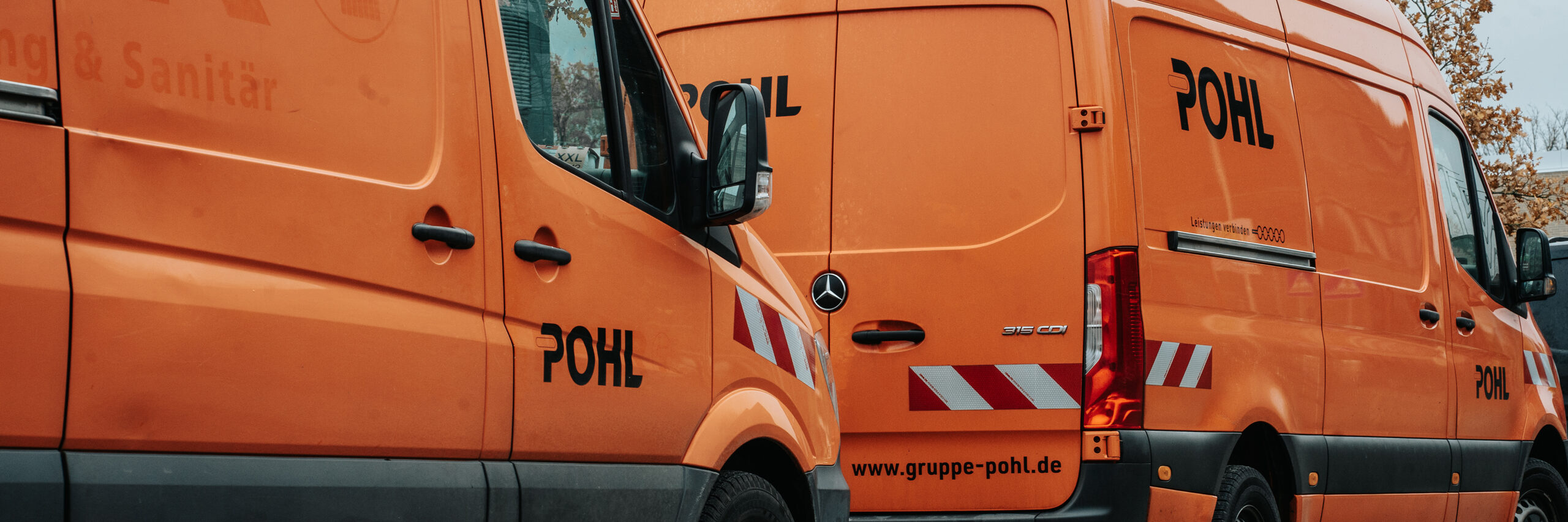 In der Mitte und rechts steht ein orangefarbenes POHL-Fahrzeug mit Heck- und Seitenansicht sowie rot-weißen Streifen; links im Bild ist die Seitenansicht eines weiteren orangefarbenen POHL-Fahrzeugs mit Außenspiegel, im Hintergrund stehen helle Bäume vor grauem Himmel.