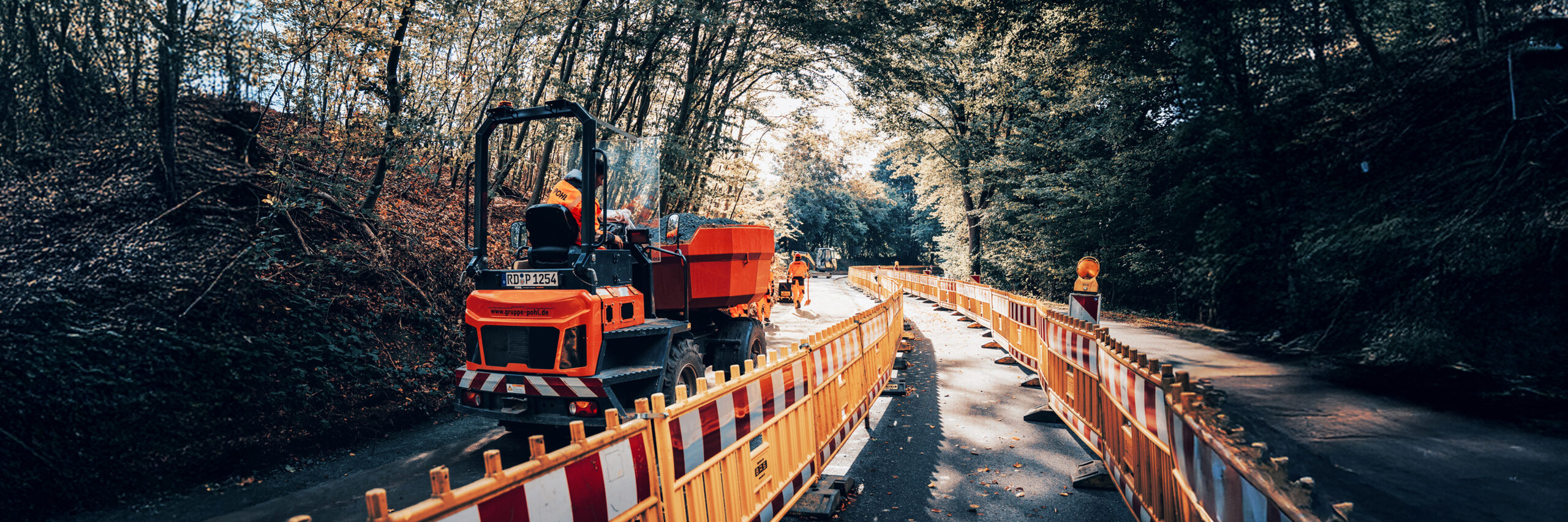 Baumaschine von POHL auf einer abgesperrten Landstraße, Mitarbeitende von POHL bei Straßenbauarbeiten.