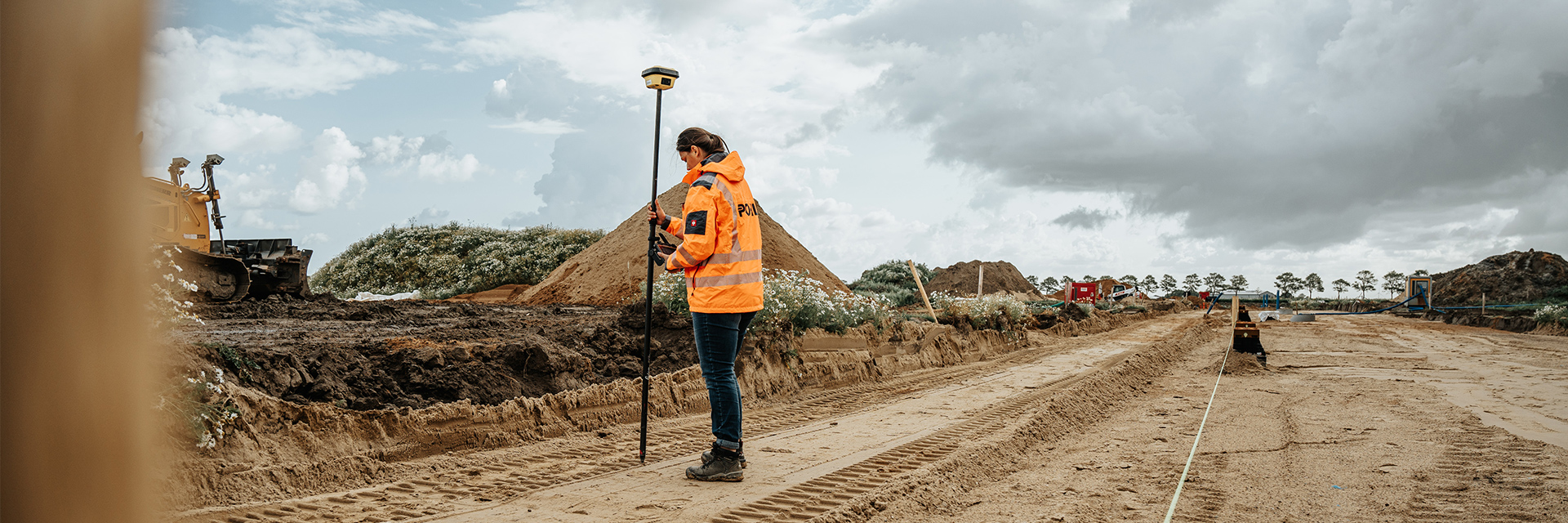 In der Mitte steht eine POHL-Mitarbeiterin in orangefarbener Jacke mit einem hohen Messstab auf einer breiten Sandtrasse; links im Hintergrund steht eine gelbe Raupe, rechts ziehen sich Erdhaufen und Bäume entlang.