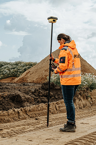 In der Mitte steht eine POHL-Mitarbeiterin in orangefarbener Jacke mit einem hohen Messstab auf einer breiten Sandtrasse; links im Hintergrund steht eine gelbe Raupe, rechts ziehen sich Erdhaufen und Bäume entlang.