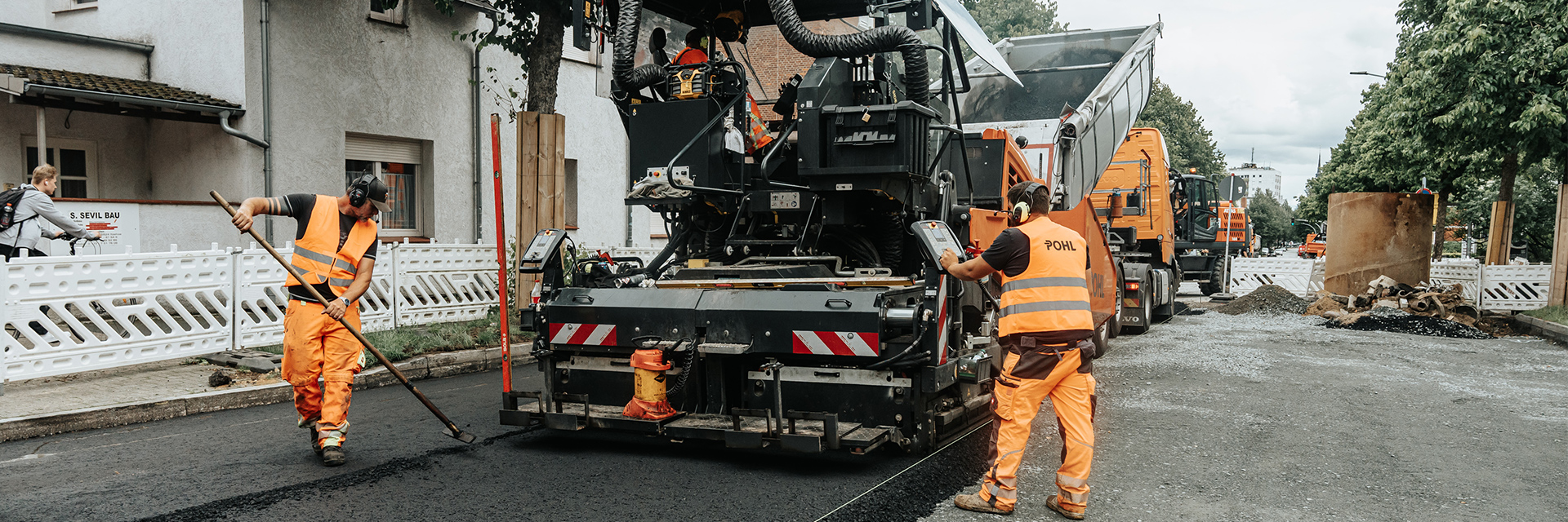 In der Mitte arbeitet eine große Straßenbaumaschine auf frischem Asphalt; links glättet ein POHL-Mitarbeiter den Belag mit einem Werkzeug, rechts bedient ein weiterer POHL-Mitarbeiter die Maschine, im Hintergrund stehen mehrere orangefarbene POHL-Fahrzeuge.