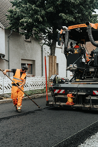 In der Mitte arbeitet eine große Straßenbaumaschine auf frischem Asphalt; links glättet ein POHL-Mitarbeiter den Belag mit einem Werkzeug, rechts bedient ein weiterer POHL-Mitarbeiter die Maschine, im Hintergrund stehen mehrere orangefarbene POHL-Fahrzeuge.