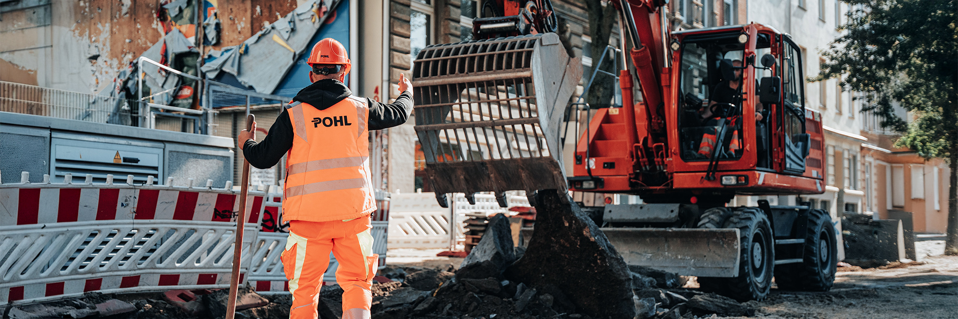 In der Mitte steht ein POHL-Mitarbeiter in orangefarbener Arbeitskleidung und mit orangefarbenem Helm mit dem Rücken zur Kamera. Rechts im Bild arbeitet eine orangefarbene POHL-Maschine mit großer Schaufel über gebrochenem Asphalt. Links im Bild stehen rot-weiße Absperrungen. Im Hintergrund liegen Hausfassaden mit Fenstern und Türen. Vorne im Bild liegen dunkle Asphaltstücke auf dem Boden.