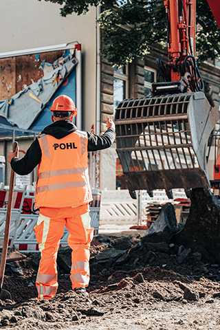 In der Mitte steht ein POHL-Mitarbeiter in orangefarbener Arbeitskleidung und mit orangefarbenem Helm mit dem Rücken zur Kamera. Rechts im Bild arbeitet eine orangefarbene POHL-Maschine mit großer Schaufel über gebrochenem Asphalt. Links im Bild stehen rot-weiße Absperrungen. Im Hintergrund liegen Hausfassaden mit Fenstern und Türen. Vorne im Bild liegen dunkle Asphaltstücke auf dem Boden.