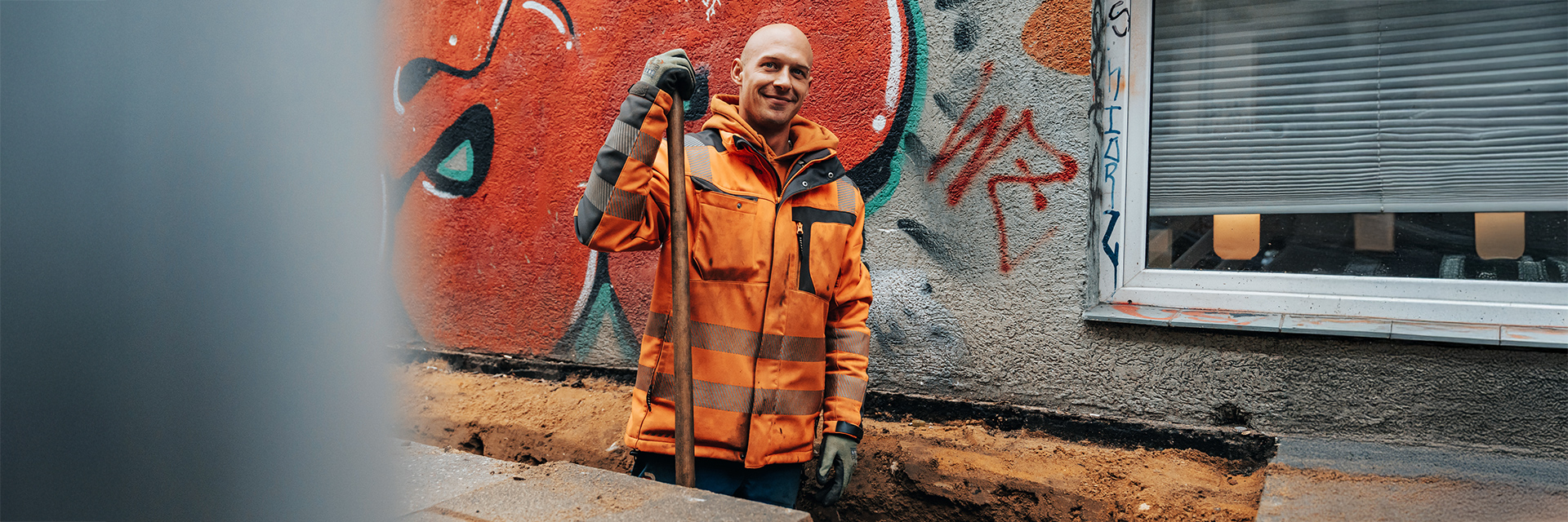In der Mitte steht ein POHL-Mitarbeiter in orangefarbener Jacke und hält einen Stiel in der Hand. Rechts im Bild befindet sich ein Fenster in einer grauen Wand mit roter Graffiti. Links im Bild verdeckt ein unscharfer Rand einen kleinen Teil der Szene. Vorne im Bild verläuft eine schmale Baugrube. Im Hintergrund zieht sich die strukturierte Hauswand über die gesamte Breite.