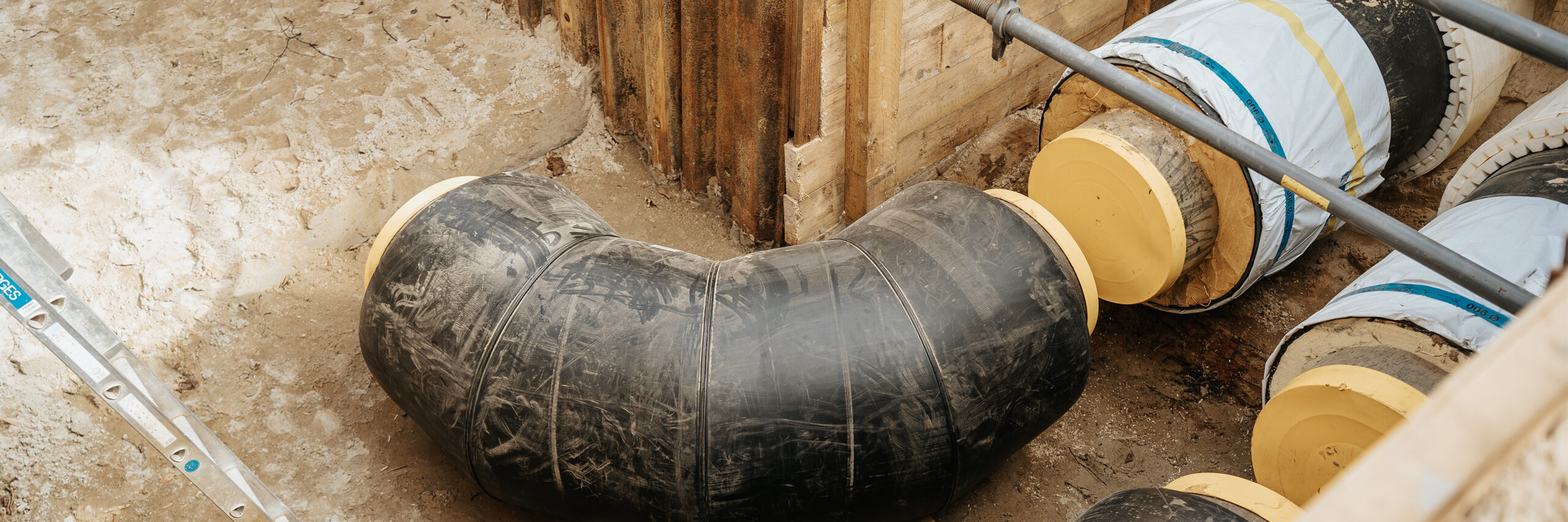 Ein Baugraben mit großen, isolierten schwarzen Rohrleitungen. Die Leitungen sind an den Enden mit gelben Kappen verschlossen und verlaufen in einer Biegung. Die Grabenwände sind mit Holz und Metallstreben abgestützt.