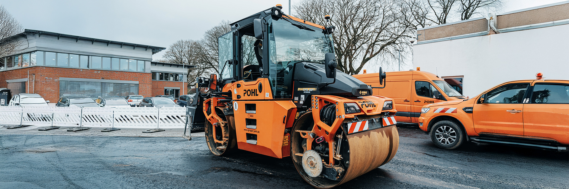 In der Mitte steht eine große orangefarbene POHL-Walze auf frisch bearbeitetem Asphalt; rechts im Bild parken orangefarbene POHL-Fahrzeuge, links stehen weiße Absperrgitter vor einem Gebäude.