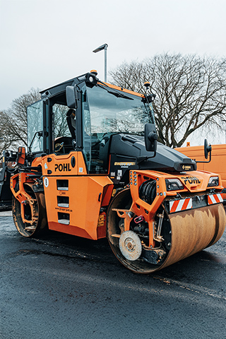 In der Mitte steht eine große orangefarbene POHL-Walze auf frisch bearbeitetem Asphalt; rechts im Bild parken orangefarbene POHL-Fahrzeuge, links stehen weiße Absperrgitter vor einem Gebäude.