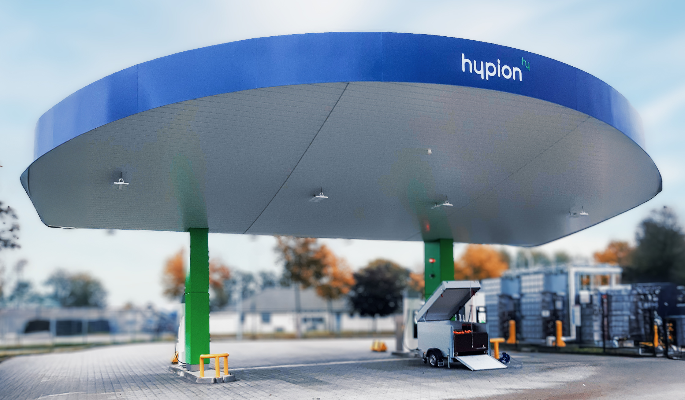 Referenz Hypion Wasserstofftankstelle Neumünster: Unten im Bild spannt sich ein rundes, blaues Tankstellendach über zwei grüne Pfeiler. Rechts daneben steht ein geöffneter Geräteanhänger; hinten sind Zäune, Bäume und Gebäude unscharf.