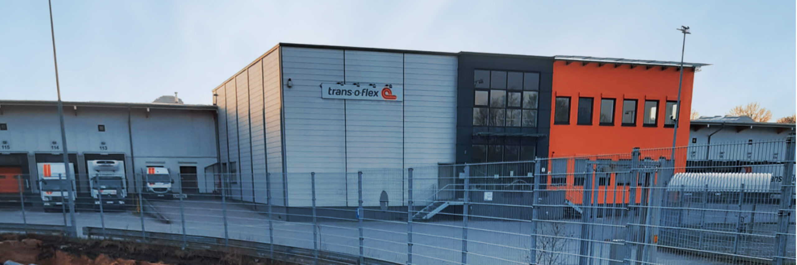 Referenz Neumünster Hypion Wasserstofftankstelle: Im Vordergrund verläuft ein langer offener Erdgraben entlang eines Metallzauns; der Boden ist mit grauer Folie ausgelegt, dahinter eine Halle mit orangefarbener Fassadenseite.