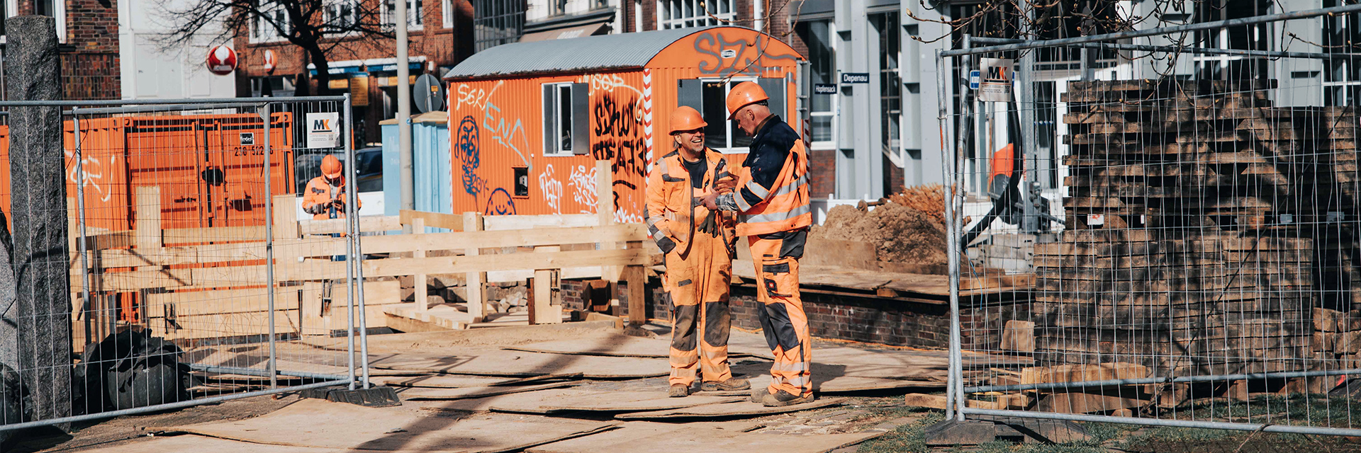 In der Mitte sprechen zwei POHL-Mitarbeiter in orangefarbener Arbeitskleidung auf einer abgesperrten Baustelle; links und rechts begrenzen Metallzäune den Bereich, im Hintergrund steht ein orangefarbener Container vor Backsteinfassaden.