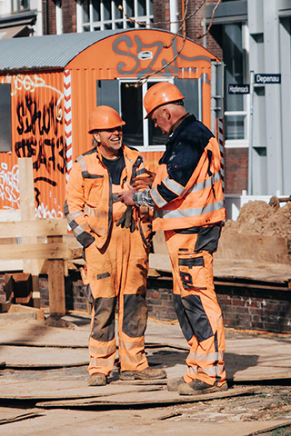In der Mitte sprechen zwei POHL-Mitarbeiter in orangefarbener Arbeitskleidung auf einer abgesperrten Baustelle; links und rechts begrenzen Metallzäune den Bereich, im Hintergrund steht ein orangefarbener Container vor Backsteinfassaden.