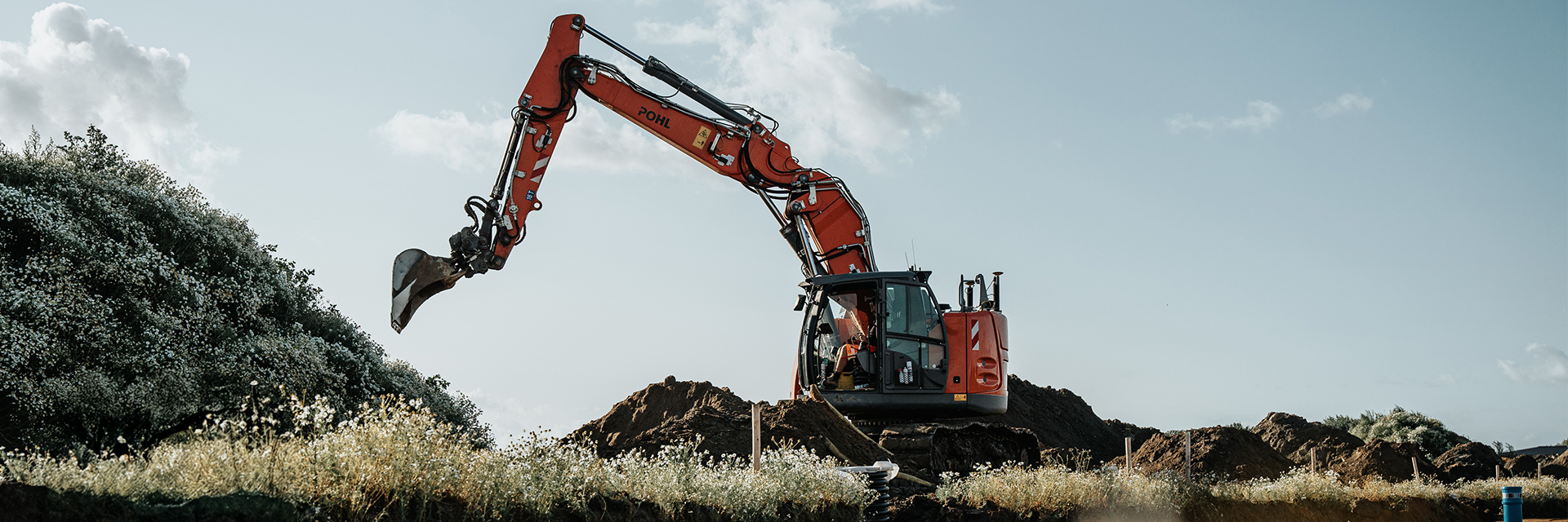 In der Mitte steht ein orangefarbener POHL-Bagger auf einem Erdhaufen; der lange Arm ragt nach links über niedrige Büsche, im Hintergrund ist blauer Himmel mit Wolken.