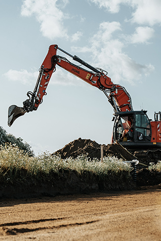 In der Mitte steht ein orangefarbener POHL-Bagger auf einem Erdhaufen; der lange Arm ragt nach links über niedrige Büsche, im Hintergrund ist blauer Himmel mit Wolken.