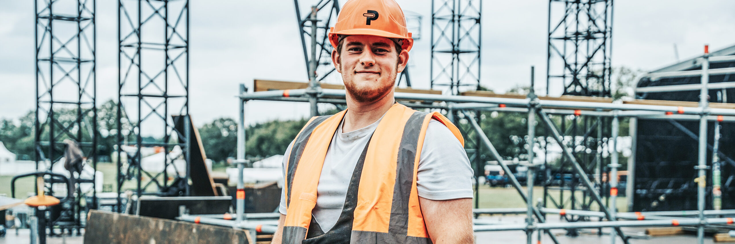 Zimmerer: In der Mitte steht ein POHL-Mitarbeiter mit orange Helm und Warnweste vor Gerüsttürmen. Links und rechts ziehen sich Metallgerüste über den Platz; der Boden ist mit Platten ausgelegt.