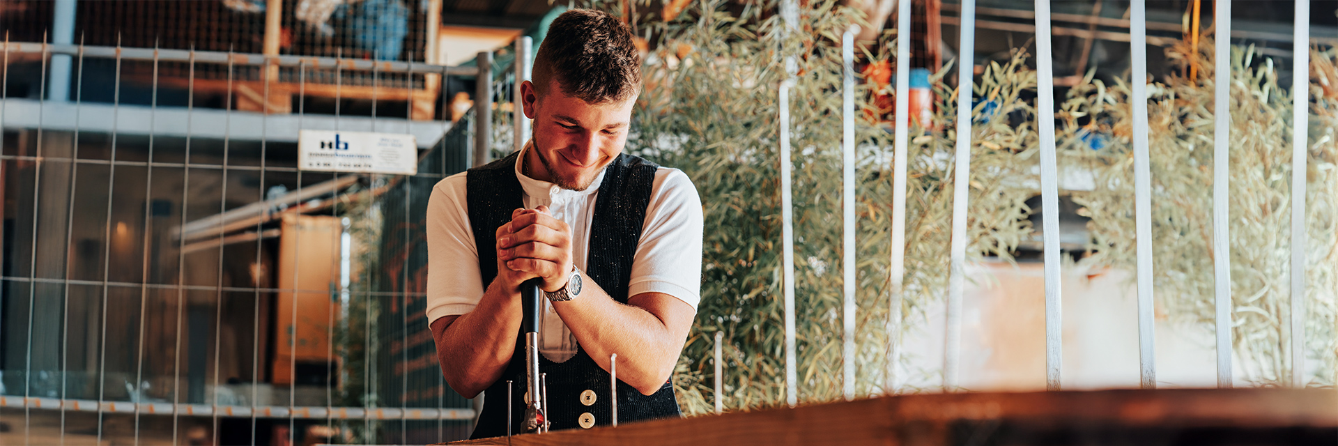 In der Mitte steht ein Mann in heller Kleidung und dunkler Weste und stützt beide Hände auf ein Werkzeug; links und rechts stehen Metallstäbe in einer Reihe, im Hintergrund sind Pflanzen und Werkstattbereiche.