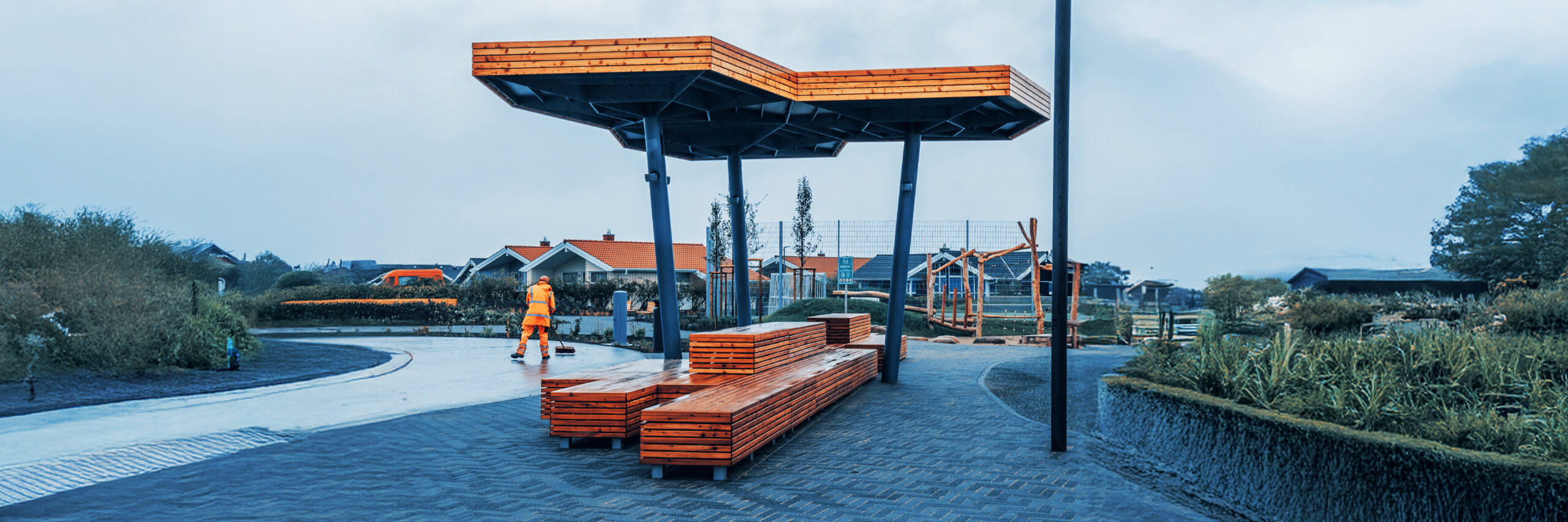 Überdachte Sitzgelegenheit mit Holzbänken in einer neu gestalteten Außenanlage, Mitarbeiter von POHL im Hintergrund.