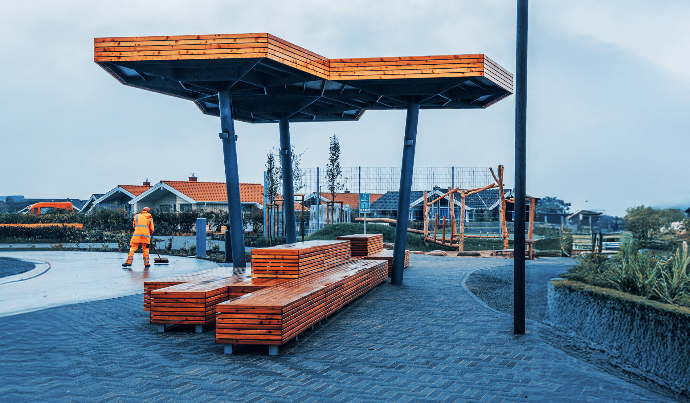 Überdachte Sitzgelegenheit mit Holzbänken in einer neu gestalteten Außenanlage, Mitarbeiter von POHL im Hintergrund.