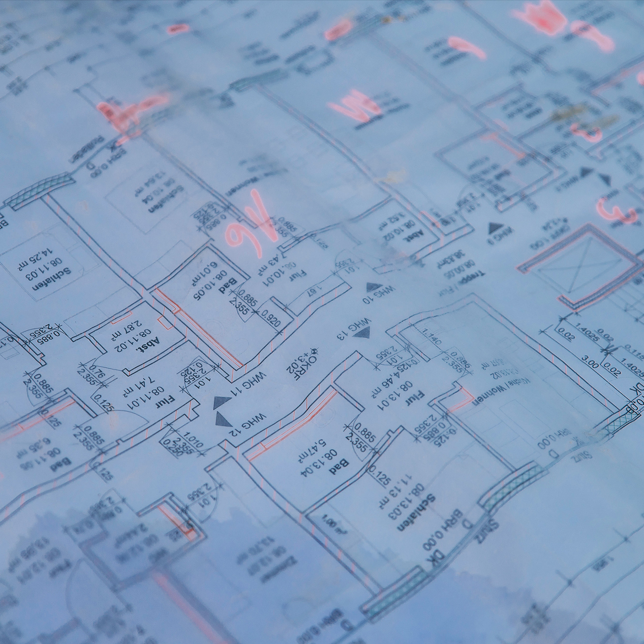 Vorne im Bild liegt ein blauer Grundrissplan mit Raumkonturen, Maßen und Pfeilen. In der Mitte markieren mehrere rosafarbene Zeichen Stellen auf dem Plan; Ränder und Faltkanten sind sichtbar.