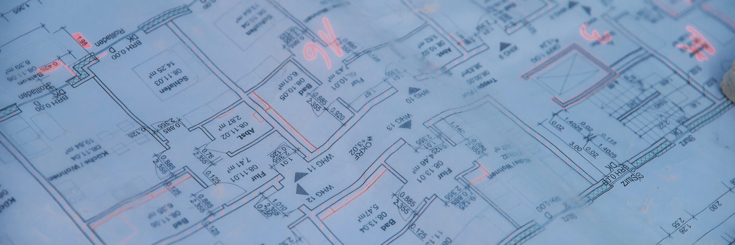 Vorne im Bild liegt ein blauer Grundrissplan mit Raumkonturen, Maßen und Pfeilen. In der Mitte markieren mehrere rosafarbene Zeichen Stellen auf dem Plan; Ränder und Faltkanten sind sichtbar.