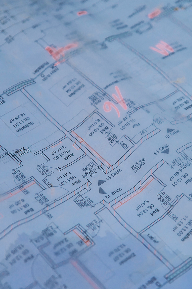 Vorne im Bild liegt ein blauer Grundrissplan mit Raumkonturen, Maßen und Pfeilen. In der Mitte markieren mehrere rosafarbene Zeichen Stellen auf dem Plan; Ränder und Faltkanten sind sichtbar.