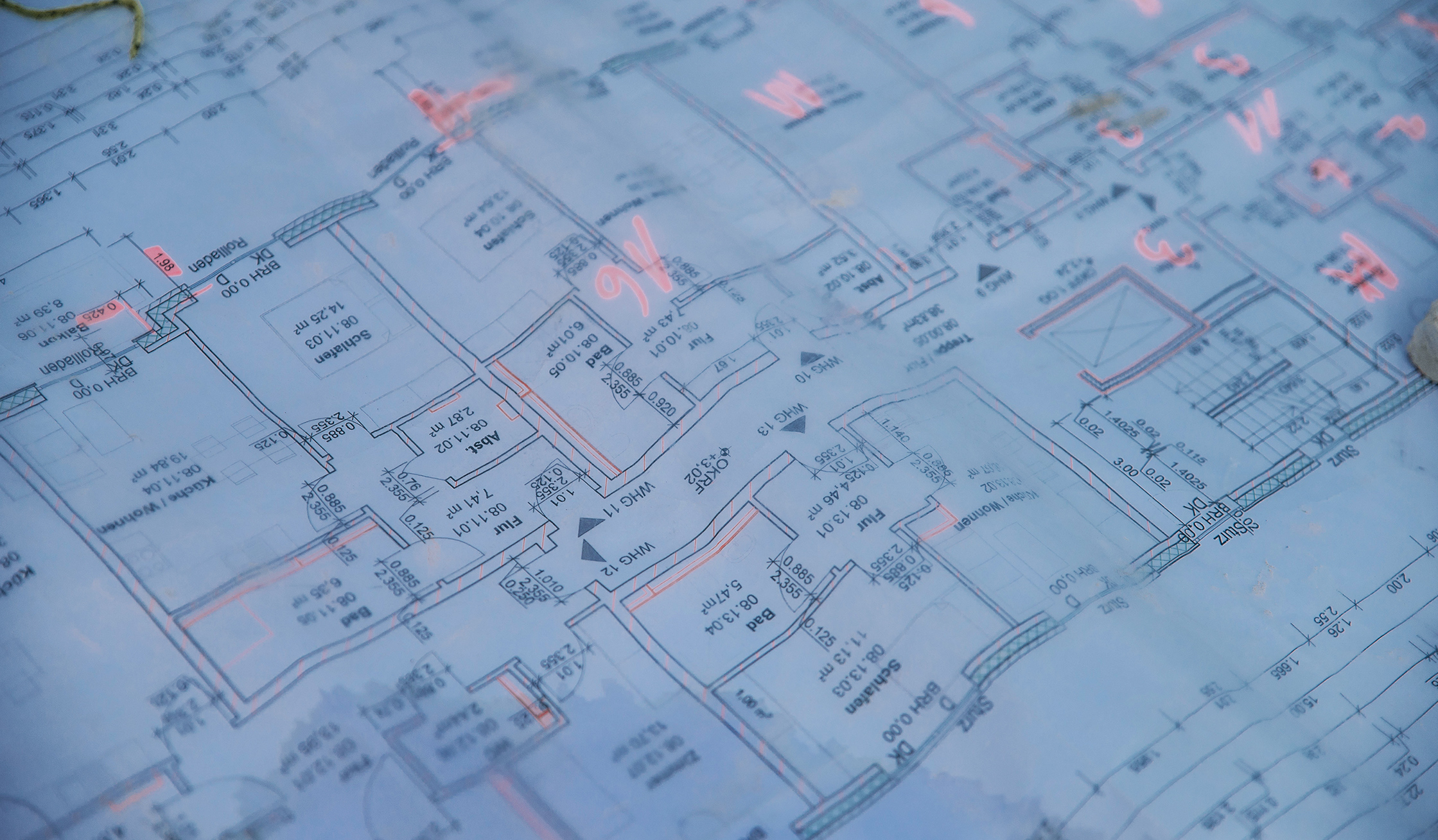Vorne im Bild liegt ein blauer Grundrissplan mit Raumkonturen, Maßen und Pfeilen. In der Mitte markieren mehrere rosafarbene Zeichen Stellen auf dem Plan; Ränder und Faltkanten sind sichtbar.